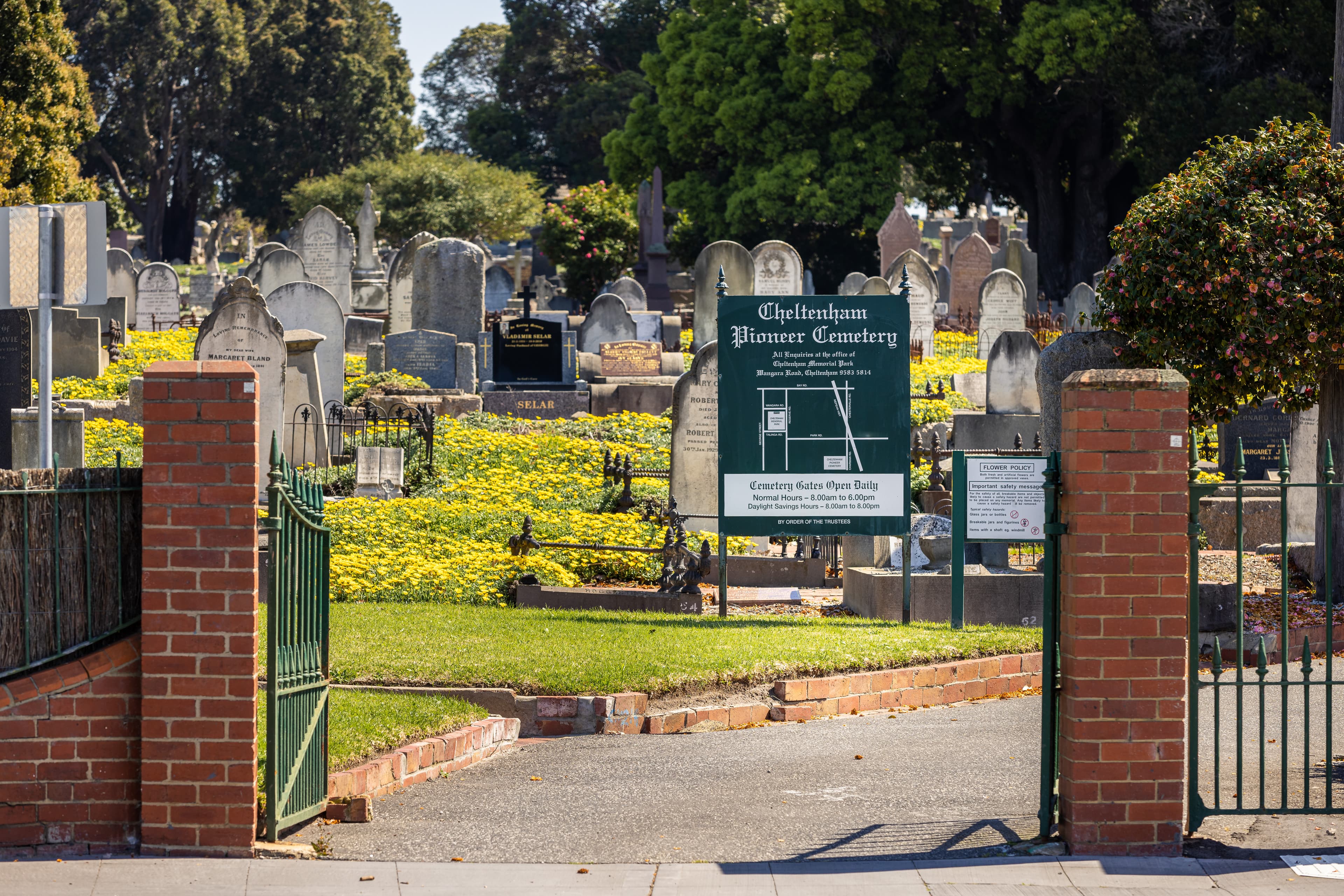 Front entrance of brick and metal gates to Cheltenham Pioneer Cemetery with a view of a map sign and gravestones in the background
