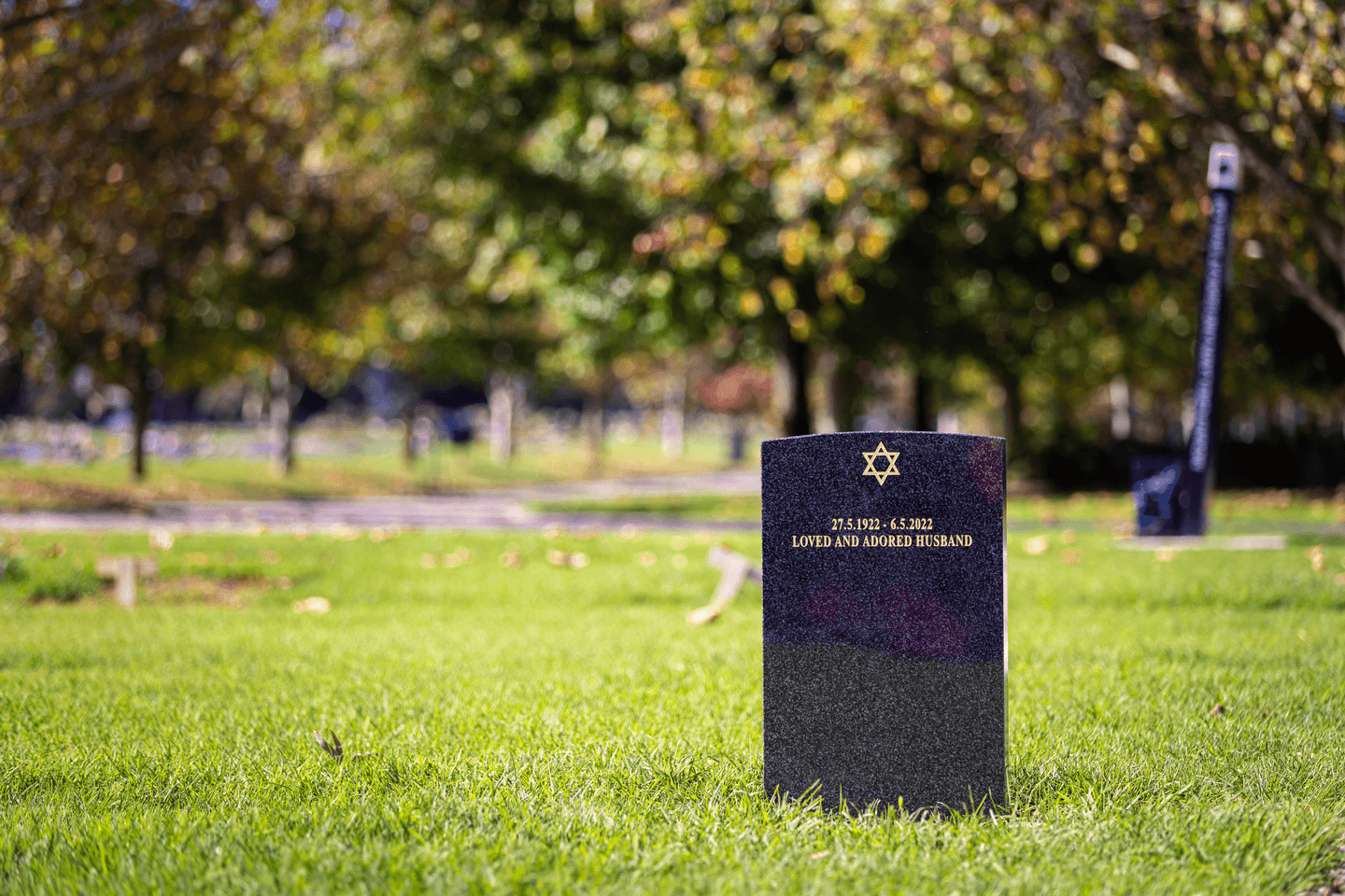 A Jewish gravestone inscribed with a Star of David, dates of birth and death, and the words "Loved and adored husband," set in a peaceful cemetery.