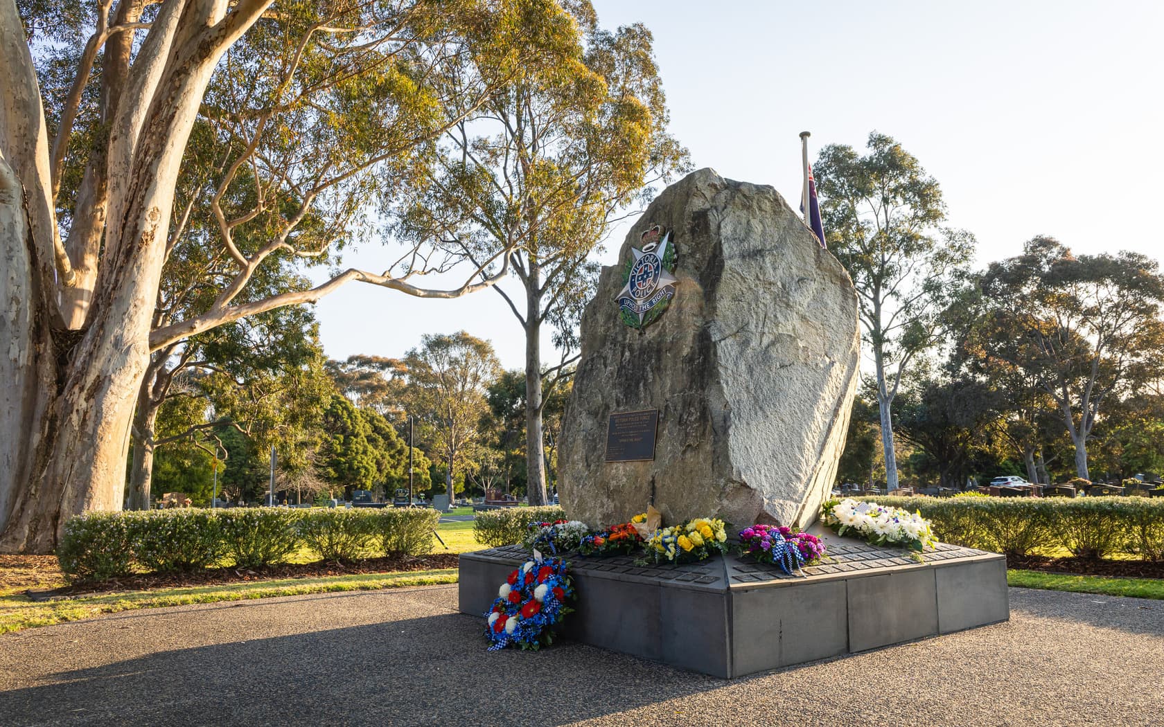 A large stone monument adorned with flowers, set in a serene park with tall trees and a clear sky.