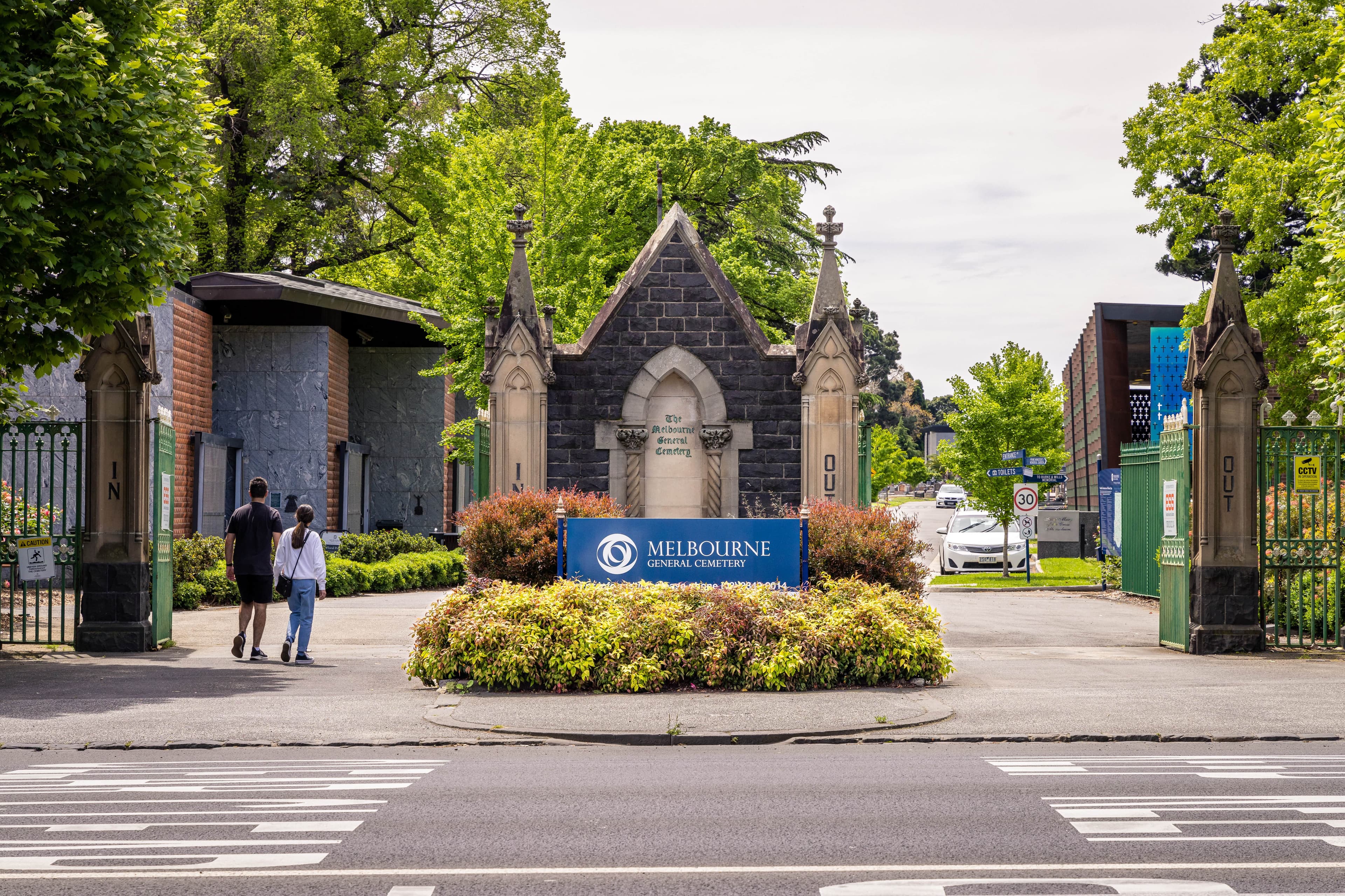 Two people walking past the entrance to Melbourne General Cemetery with a brick chapel in the centre between the gates