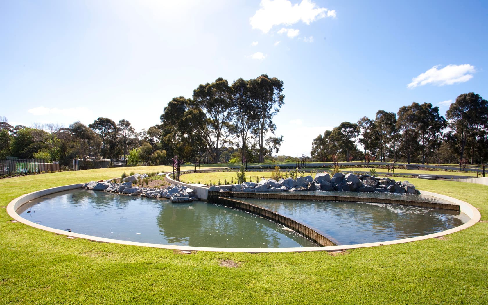 A serene area scene featuring a curved water and rock feature surrounded by green grass and trees under a clear blue sky.