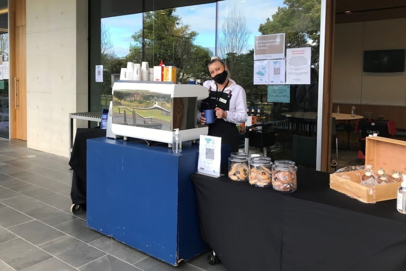 A photograph of an SMCT staff member standing behind a coffee machine set up next to a trestle table of cookies and muffins. The SMCT staff member is standing behind a coffee machine and wearing a face mask. 