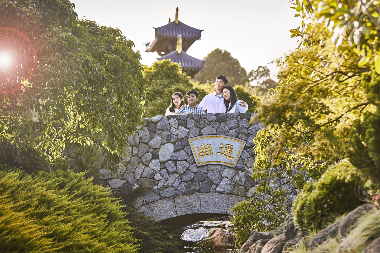 An asian family standing together on a stone bridge adorned with a golden inscription, surrounded by leafy trees and a traditional pagoda in the background.
