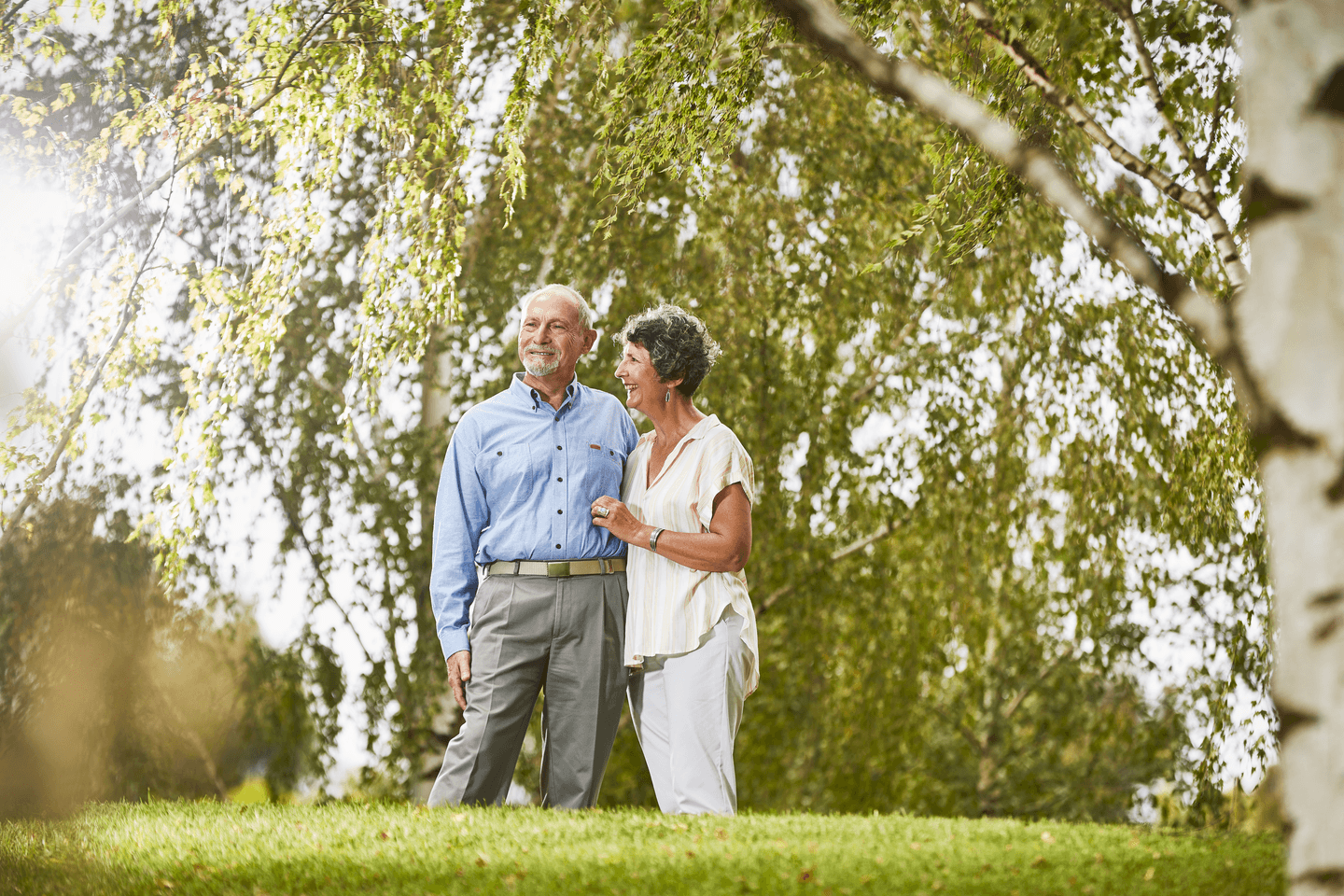 an elderly man and woman embracing each other standing in lawn beneath a large tree