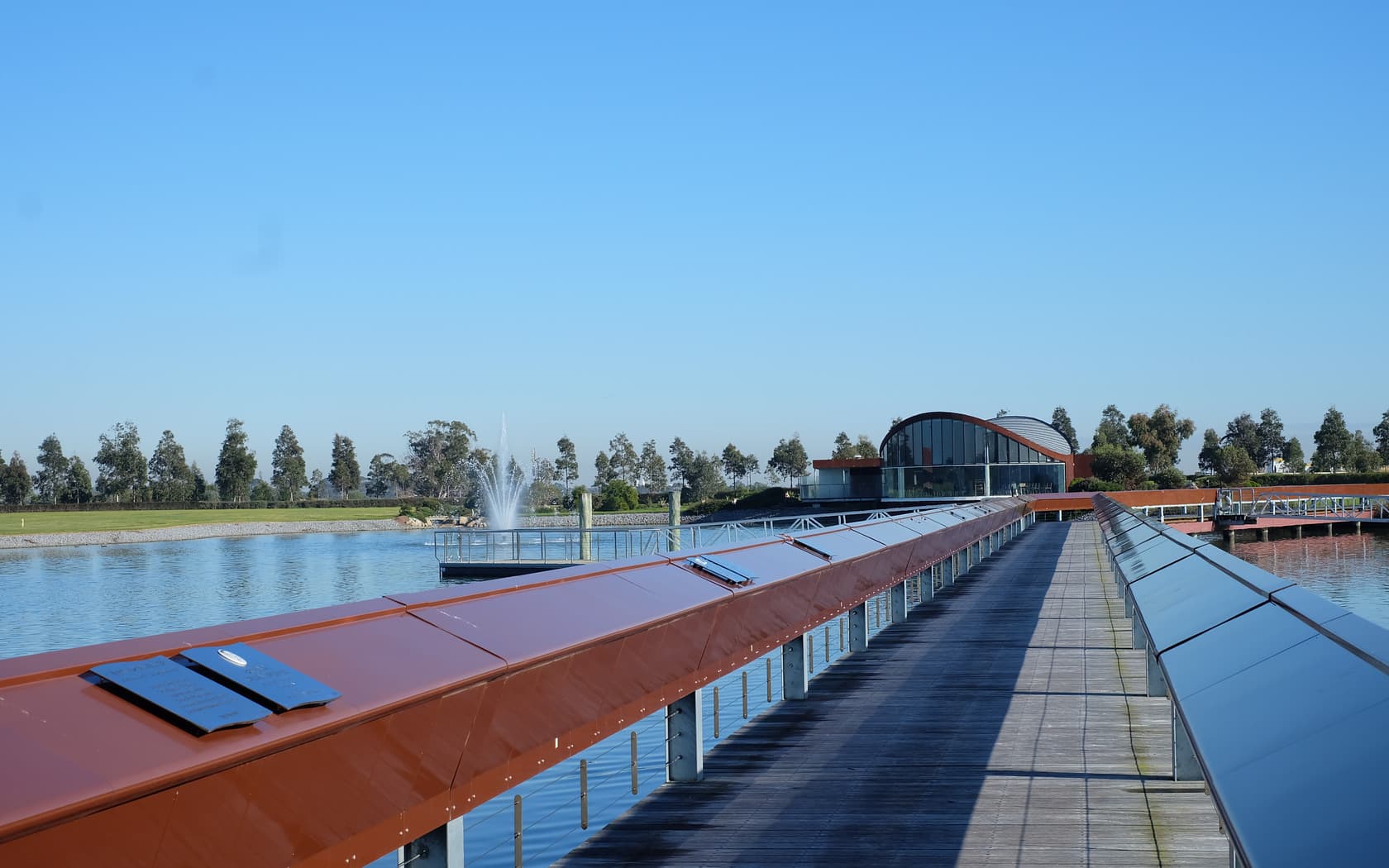 A wooden walkway with plaques extends over a calm lake, leading to a modern building, with a fountain spraying in the background under a clear blue sky.