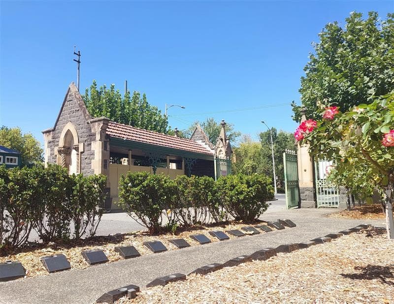 A small stone chapel with a red-tiled roof is surrounded by trees and shrubs on a sunny day. A path lined with cremation plaques leads towards the entrance.