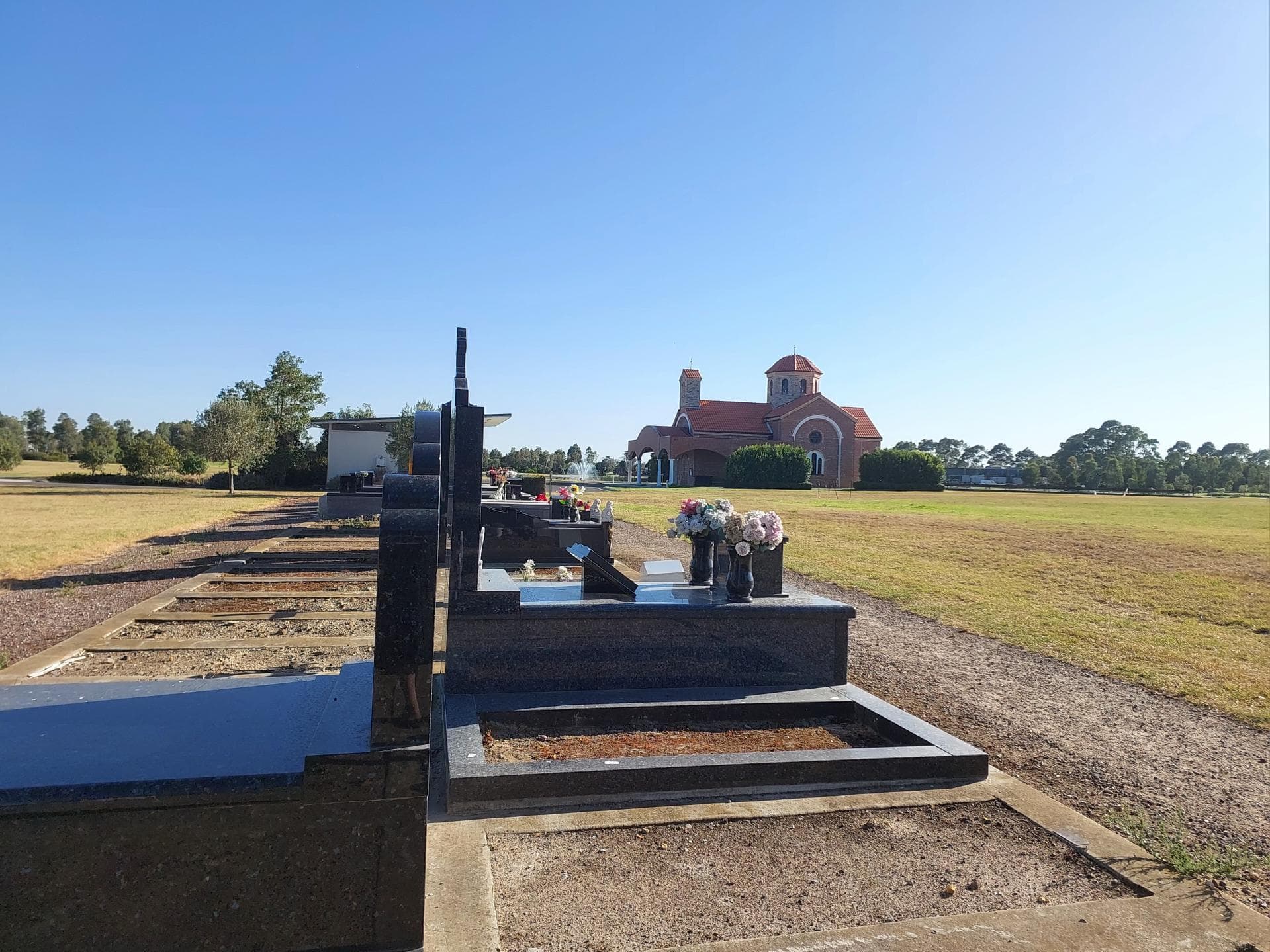 A peaceful cemetery scene featuring grand double-row monumental headstone and a chapel in the background under a clear blue sky.