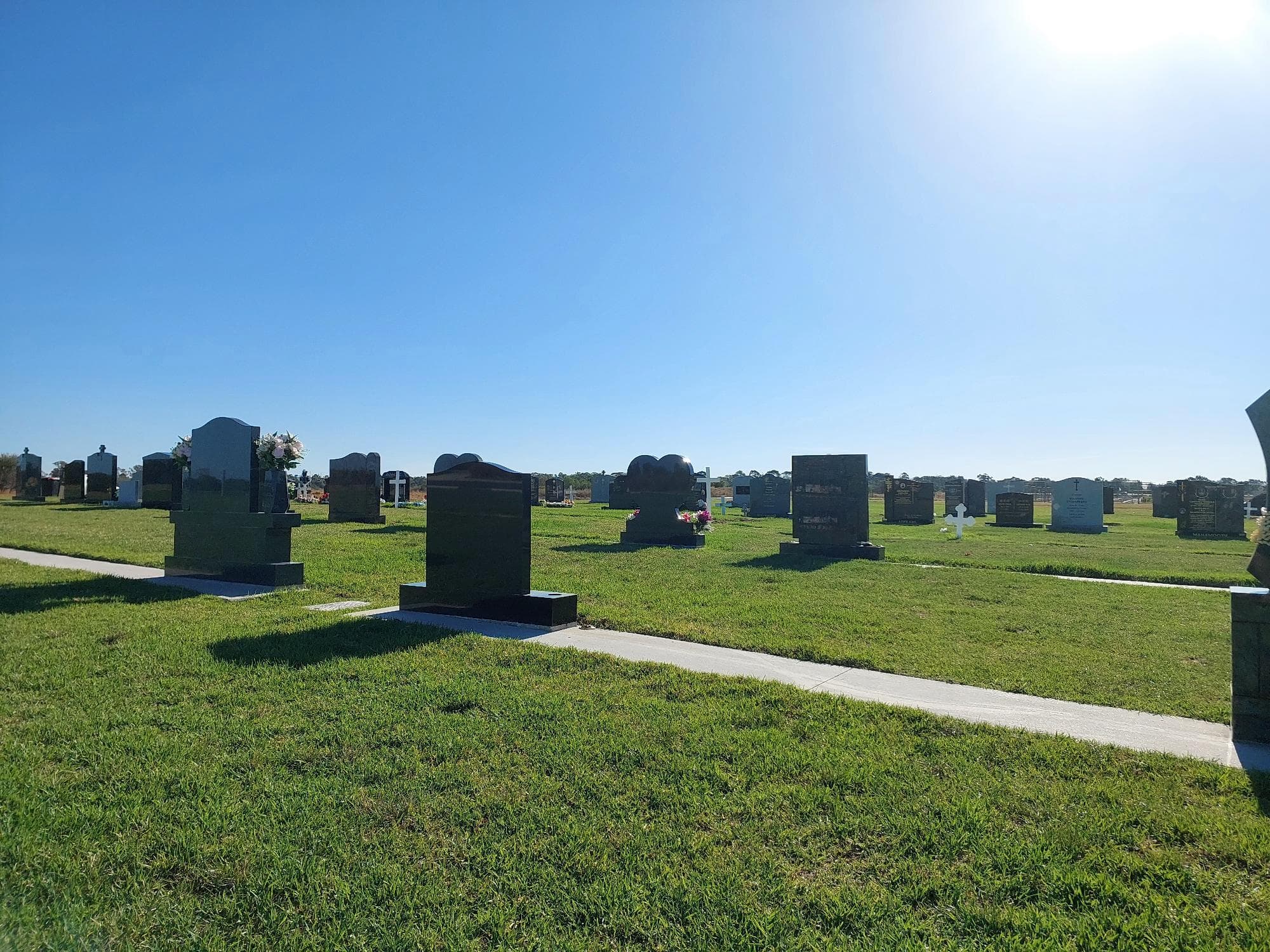 A serene cemetery scene with various gravestones on green grass, under a clear blue sky, bathed in sunlight.