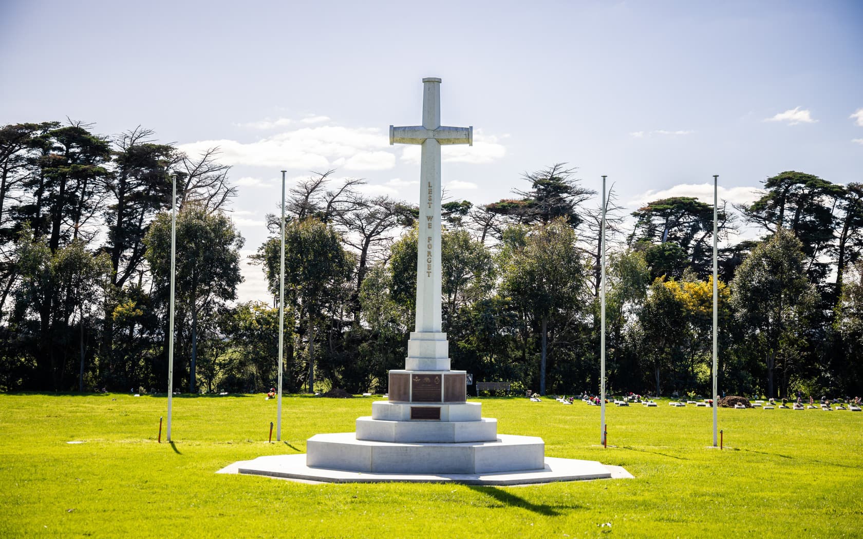 A tall white stone cross monument stands in a grassy memorial area surrounded by trees, with the inscription "Lest We Forget" visible.