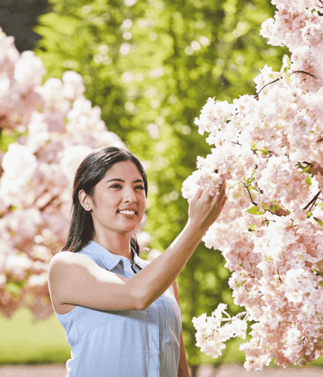 Woman in a sleeveless blouse gently touches pink cherry blossoms in a sunlit garden.